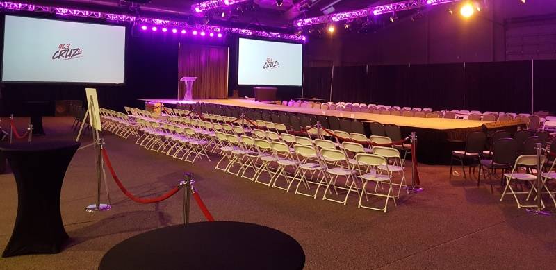 A fashion show with rows of chairs surrounding a stage and projector screens with pink tinted spotlights hanging from installed trusses